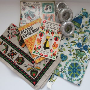Pode incluir: Dois livros de cozinha vintage da Pensilv&acirc;nia holandesa com capas amarelas e vermelhas com desenhos de arte popular. Os livros de cozinha s&atilde;o intitulados "Dutch Cookbook" e "Pennsylvania Dutch Cookbook."  Os livros de cozinha s&atilde;o cercados por uma toalha de ch&aacute; de linho branco com um design de arte popular preto, verde e vermelho, uma toalha de ch&aacute; floral azul e branca e tr&ecirc;s an&eacute;is de bolo de prata.