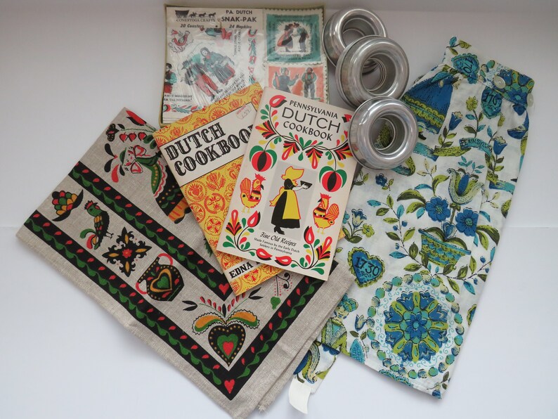 Pode incluir: Dois livros de cozinha vintage da Pensilv&acirc;nia holandesa com capas amarelas e vermelhas com desenhos de arte popular. Os livros de cozinha s&atilde;o intitulados "Dutch Cookbook" e "Pennsylvania Dutch Cookbook."  Os livros de cozinha s&atilde;o cercados por uma toalha de ch&aacute; de linho branco com um design de arte popular preto, verde e vermelho, uma toalha de ch&aacute; floral azul e branca e tr&ecirc;s an&eacute;is de bolo de prata.