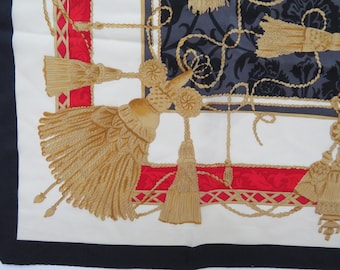 May include: Silk scarf with a detailed design featuring gold tassels, rope, and floral patterns. The scarf has a white background with red and black borders. The design includes a dark gray floral pattern.