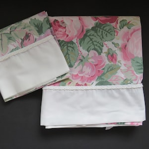 May include: Two white pillowcases with a floral print of pink roses and green leaves. The pillowcases have a white lace trim.