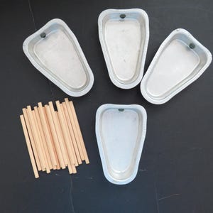 May include: Four vintage aluminum ice pop molds with a small hole at the top, and a bundle of wooden sticks. The molds have a unique shape, and are arranged on a dark surface. A classic set for making homemade frozen treats.