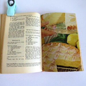 Puede incluir: Libro de cocina abierto con una receta de pastel de melocot&oacute;n y ciruela y una foto de un pastel de merengue de lim&oacute;n con una corteza de celos&iacute;a. La receta incluye ingredientes como melocotones en rodajas, ciruelas rojas, az&uacute;car, harina y mantequilla. El pastel es de un hermoso color amarillo dorado con una cobertura de merengue blanco.