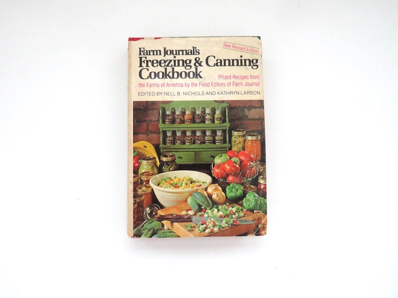 May include: A hardcover cookbook titled Farm Journal's Freezing & Canning Cookbook, edited by Nell B. Nichols and Kathryn Larson. The cover features a green spice rack, jars of preserved food, a bowl of vegetables, and chopped produce on a board.
