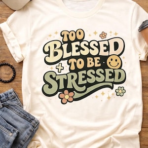 Könnte beinhalten: Cremefarbenes T-Shirt mit dem Schriftzug "TOO BLESSED TO BE STRESSED" in einem Retro-Schriftstil. Das Design umfasst ein Smiley, Blumen und Sterne. Ein lässiges, bequemes T-Shirt für den Alltag.