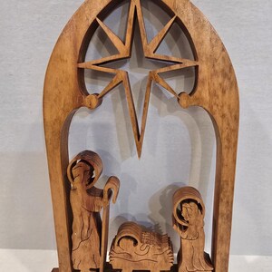 May include: Wooden nativity scene featuring a star, two figures, and a manger. The figures are carved in detail, and the wood has a warm, natural finish. The star is at the top, and the scene is set within an arched frame.
