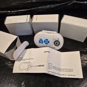 Könnte beinhalten: Ein weißer, ovaler Schrittzähler mit einem kleinen integrierten Kompass, blauen medizinischen Logos sowie dem Text "PRESS" und "LIFT" mit Pfeilen. Er wird von drei weißen Pappschachteln und einer Bedienungsanleitung mit Text begleitet.