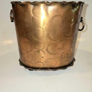 May include: A copper-colored metal bucket featuring a scalloped top and bottom edge. The exterior surface is decorated with a repeating pattern of circular indentations and includes two small metal ring handles attached to the sides of the vessel.