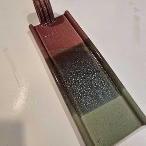 Puede incluir: Un colgante rectangular con un lazo en la parte superior. El colgante presenta un degradado de colores, comenzando con un marrón rojizo, pasando a un gris oscuro moteado y terminando con un verde claro. Los bordes están ligeramente elevados.