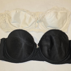 May include: A strapless bra set consisting of one white lace bra and one black smooth fabric bra. Both bras feature molded cups, underwire support, and a small decorative charm centered between the cups. The white bra has lace side panels.