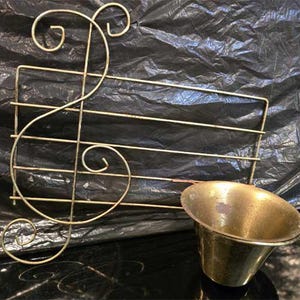 Puede incluir: Decoración de pared dorada con temática musical. El diseño presenta una clave de sol y barras horizontales, con un pequeño cuenco de latón. La decoración es probablemente para exhibir partituras o objetos pequeños.