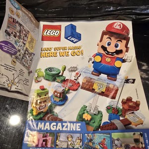 May include: A LEGO Life magazine featuring Super Mario on the cover. The cover displays a LEGO Mario figure, various Super Mario characters, and building sets. Text includes LEGO Life, LEGO Super Mario Here We Go!, and The Magazine. The bottom shows small panels labeled New LEGO Models, Comic Action, Activities, and Cute Animals.