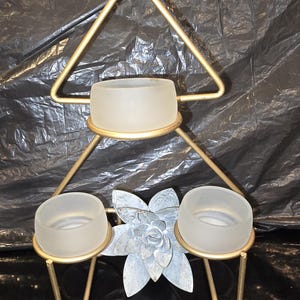 May include: A gold-toned metal stand featuring a triangular frame with three circular holders. Each holder contains a small, round, frosted glass bowl. A decorative, silver-colored metal flower is positioned centrally between the two lower bowls.