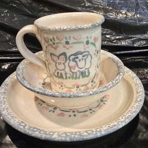 Könnte beinhalten: Ein Keramik-Tassen-, Schüssel- und Teller-Set. Die cremefarbenen Teile haben einen blauen Rand und sind mit rosa Blumen und grünen Blättern verziert. Die Tasse zeigt ein Kaninchen- und Schaf-Design.