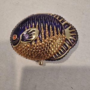 Op de afbeelding: Een decoratieve, visvormige sieradendoos. De doos is voornamelijk marineblauw met gouden accenten. De onderste helft is bedekt met kleine, goudkleurige strass-steentjes. De vis heeft een rood oog en een gouden sluiting.