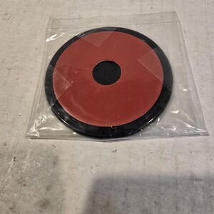 May include: A circular black plastic disc with a red adhesive backing, featuring a central circular cutout. The object is enclosed in a clear, thin plastic protective sleeve, showcasing the contrast between the black outer rim and the red center.