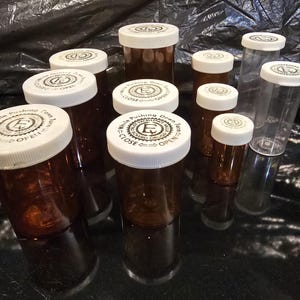 Puede incluir: Una colección de frascos de pastillas de plástico marrón y transparente con tapas blancas. Las tapas tienen el texto "While Pushing Down Turn", "Open" y "Close". Los frascos son de varios tamaños y están sobre una superficie negra reflectante.