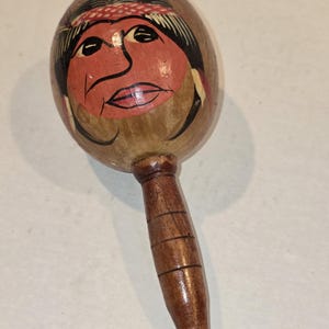 May include: A wooden maraca with a painted face design. The face is painted with red, black, and tan colors, and features a red headband. The handle is a dark brown color.