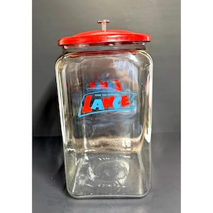 May include: A clear glass jar with a red lid and a silver handle. The jar is rectangular and features the text "FROM THE HOUSE OF LANCE" in red and blue lettering. The jar is empty.