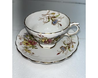 May include: A white teacup and saucer set with a floral design. The cup and saucer have a scalloped edge and gold trim. The floral pattern includes pink, yellow, and blue flowers.