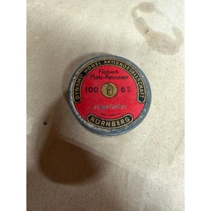 May include: A round, metal tin with a red center and black and white text. The text includes "Dynamit Nobel Aktiengesellschaft" and "Nurnberg". The center of the tin has the text "100" and "6", with an acorn graphic.