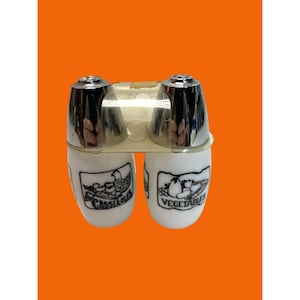 Könnte beinhalten: Ein Vintage-Salz- und Pfefferstreuer-Set. Das Set enthält zwei weiße Keramikstreuer mit schwarzen Illustrationen und Text. Die Oberteile sind verchromt. Auf einem Streuer steht "Casseroles" und auf dem anderen "Vegetables".