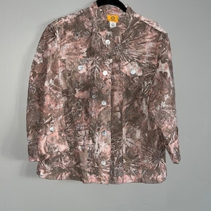 May include: A button-down shirt with a floral pattern in shades of brown and pink. The shirt has two chest pockets and white buttons. The collar has a small gold-colored emblem.