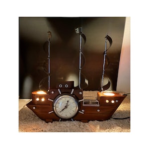 May include: A wooden ship-shaped decorative clock with a vintage aesthetic. The clock features a round face with Roman numerals and is flanked by two candle holders. The ship has metal sails and is lit with warm light.