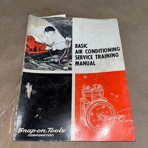 May include: A vintage Snap-on Tools Basic Air Conditioning Service Training Manual. The cover features a black and white illustration of a mechanic working on a red air conditioning unit, with the title in bold black letters. The lower left corner has the Snap-on Tools Corporation logo.