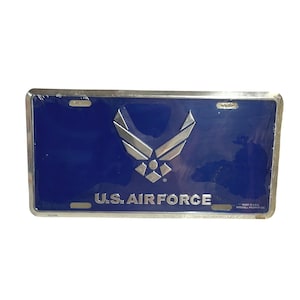 May include: A blue license plate with a silver emblem and the text "U.S. AIR FORCE." The plate has a silver border and is wrapped in clear plastic. The emblem is a stylized design of a bird or wings.