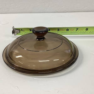 May include: A round, transparent brown glass lid with a central knob handle. A yellow measuring tape is positioned behind the lid, indicating a diameter of approximately 8 inches across the circular glass surface.