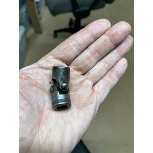 May include: A used, metal universal joint socket wrench. The tool is a dark grey colour, showing signs of wear. It has a square socket end and a ball-and-socket joint for flexible use. The tool is held in a hand.