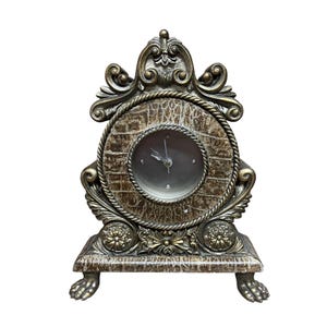 May include: An antique-style clock with a brown, textured face and ornate gold-colored frame. The clock features a round face with black hands and is set in a decorative, detailed base with claw feet. The clock's design includes rope-like and floral accents.