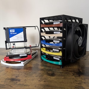 Puede incluir: Un bastidor negro para discos duros de ordenador con un ventilador. El bastidor contiene varios discos duros, cada uno separado por divisores de colores. También se ve un SSD SATA WD Blue 500GB 3D NAND. El bastidor está sobre una superficie de madera.