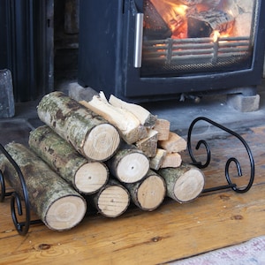 May include: A black metal log rack with a decorative scroll design holds a stack of cut logs. The logs are arranged in a staggered pattern and have a natural bark finish.