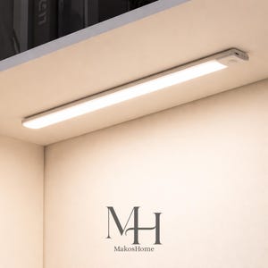 Op de afbeelding: Een heldere, rechthoekige LED-lichtarmatuur, geïnstalleerd onder een plank. Het licht straalt een warme, witte gloed uit. De armatuur is gemonteerd op een wit oppervlak. Het logo "MH MakosHome" is onderaan zichtbaar.