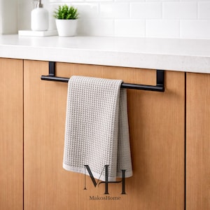 Può includere: Un portasciugamani in metallo nero montato su una porta di un mobile in legno, con un asciugamano a nido d'ape grigio chiaro. Il portasciugamani ha un design semplice e moderno. Il marchio "MakosHome" è visibile in basso.