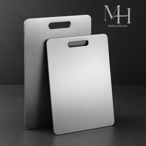 May include: Two rectangular, stainless steel chopping boards with a brushed finish. Each board has a rectangular handle cutout at the top. The boards are leaning against each other on a dark background. The MakosHome logo is in the upper right corner.
