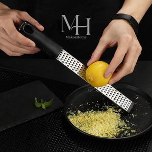Könnte beinhalten: Ein Edelstahl-Reibeisen mit schwarzem Griff wird verwendet, um eine gelbe Zitrone zu schälen. Die Schale fällt auf einen schwarzen Teller, mit einem kleinen Zweig grüner Blätter auf einer schwarzen Oberfläche. Das Logo "MH MakosHome" ist sichtbar.