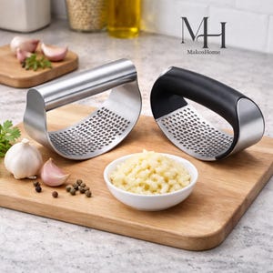 Könnte beinhalten: Zwei Knoblauchpressen, eine mit schwarzem Griff und eine mit silbernem Griff, liegen auf einem Holzschneidebrett. Eine kleine Schüssel mit gehacktem Knoblauch, ganzen Knoblauchzehen und Pfefferkörnern befindet sich ebenfalls auf dem Brett. Das Logo "MakosHome" ist im Hintergrund.