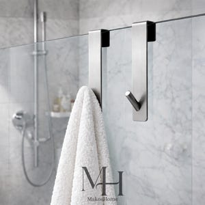 May include: Two brushed steel shower door hooks with a white towel hanging on one. The hooks are designed to hang over the glass shower door. The background shows a shower head and marble tile.