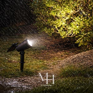 Puede incluir: Un foco negro ilumina una escena de jardín, con lluvia cayendo. La luz brilla sobre la hierba y un arbusto. El foco está montado sobre una estaca negra. El texto "MH MakosHome" está en la parte inferior.