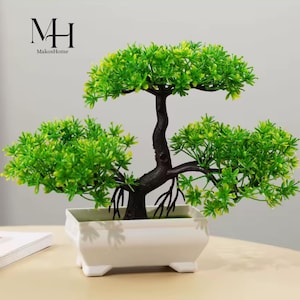 Può includere: Un piccolo bonsai artificiale in un vaso rettangolare bianco. L'albero ha un tronco e rami marrone scuro con un fogliame verde brillante. Il vaso è quadrato con bordi arrotondati e poggia su quattro piedini. Il logo "MH MakosHome" è nell'angolo in alto a sinistra.
