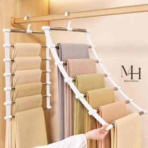May include: A white multi-tier pants hanger with several pairs of folded pants in various colors, including beige, pink, and green. The hanger is suspended from a wooden rod in a closet. The brand name "MakosHome" is visible.