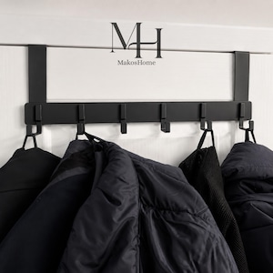 May include: A black metal over-the-door coat rack with six hooks, holding dark jackets. The rack has the text "MakosHome" and the letters "MH" on the top. The hooks are holding several coats.