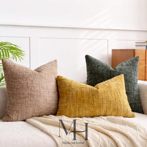 Puede incluir: Tres cojines decorativos texturizados en tonos beige, amarillo mostaza y verde oscuro descansan sobre un sofá blanco. Los cojines tienen un patrón tejido y están dispuestos con una manta color crema. El logotipo "MakosHome" es visible.
