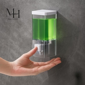Pode incluir: Um dispensador de sabonete transparente montado na parede com uma tampa branca e um botão de pressão. O dispensador é preenchido com sabonete líquido verde. A marca "MakosHome" é visível no canto superior esquerdo.