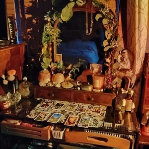 May include: A wooden vanity with a mirror, decorated with greenery and candles. Tarot cards are laid out on the surface. The vanity also features small decorative jars, bottles, and a small statue.
