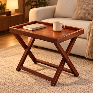 Puede incluir: Una mesa bandeja rectangular de madera con acabado marrón oscuro y borde elevado. La mesa cuenta con una base plegable en forma de X. Un libro beige cerrado y una taza de cerámica color crema con relieve están sobre la superficie plana.