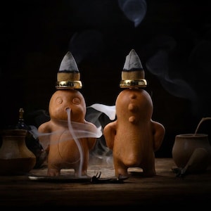 May include: Two light brown, clay-like creature figurines standing side-by-side. Each figure has a gold-colored ring on its head supporting a burning incense cone. White smoke flows from the mouth of the left figure and around both bodies.