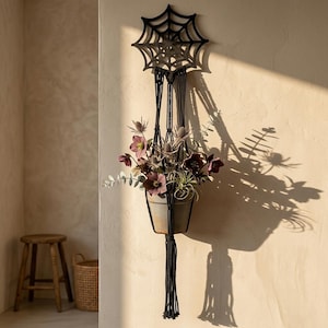 Op de afbeelding: Een zwarte macramé plantenhanger met een spinnenwebontwerp aan de bovenkant, met een terracotta pot gevuld met een bloemstuk van donkerpaarse nieskruidbloemen, stekelige distels en groene eucalyptusbladeren, afgewerkt met een lange kwast.